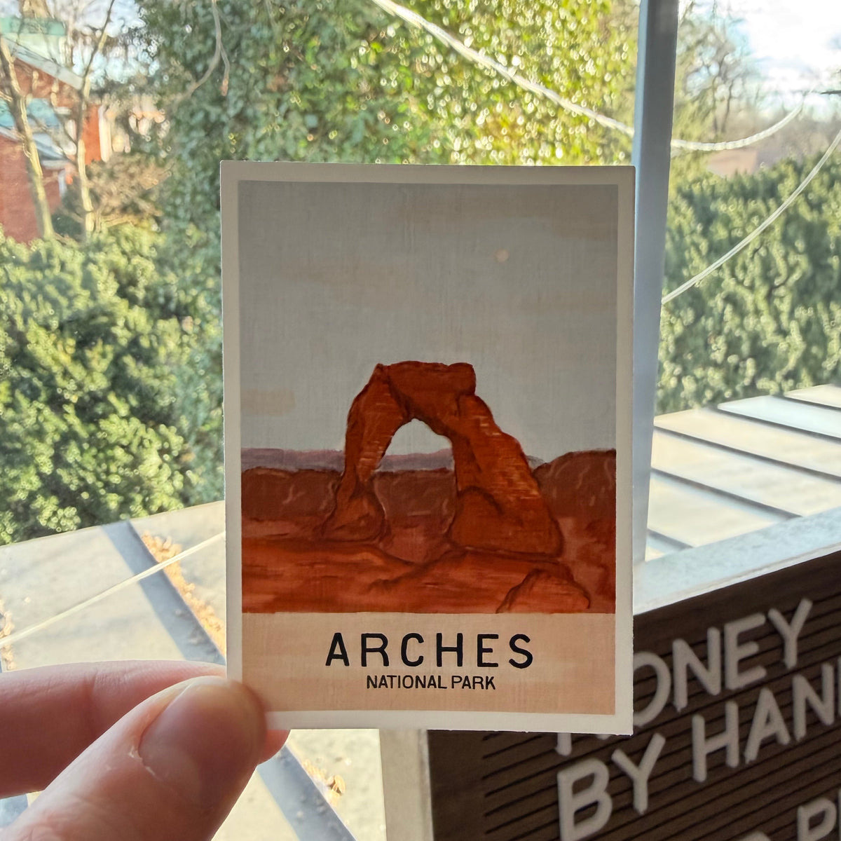 Card with Arches National Park design held up to a window with a view of trees and buildings.