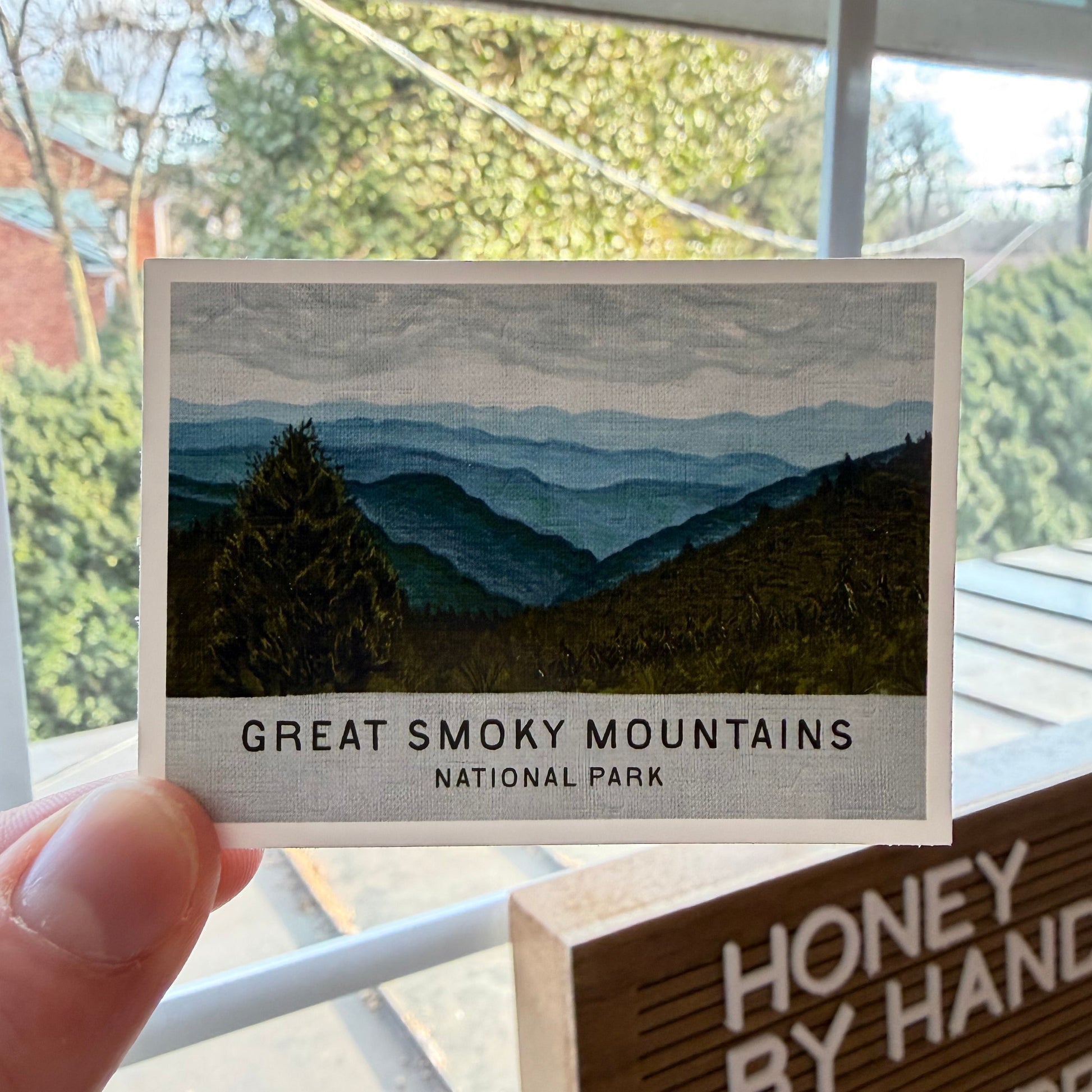 Card with Great Smoky Mountains National Park design held by a hand, with a window view in the background.